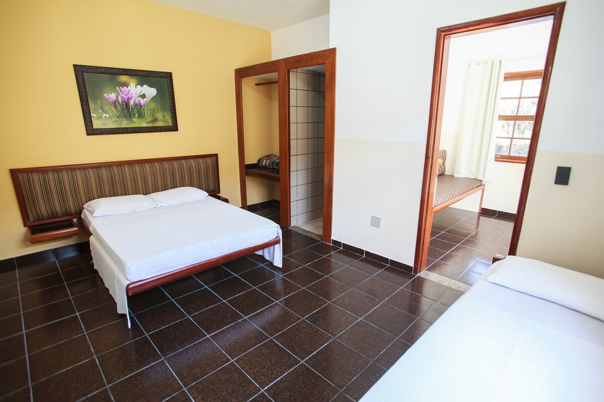 A hotel room with a bed, a wooden headboard, a tiled floor, a painting on the wall, and a door leading to another room.