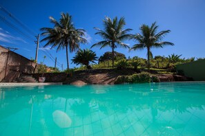 A swimming pool with clear blue water, surrounded by palm trees and a stone wall. - Guarapousada (Guarapari)