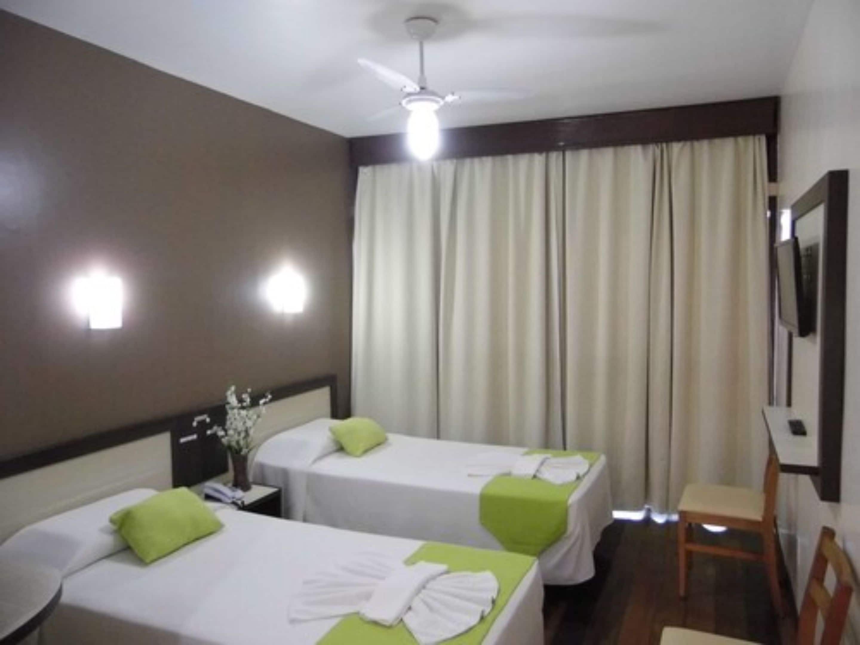 A hotel room with two beds, each with a green pillow and white linens, a ceiling fan, a TV mounted on the wall, and a large window with curtains.