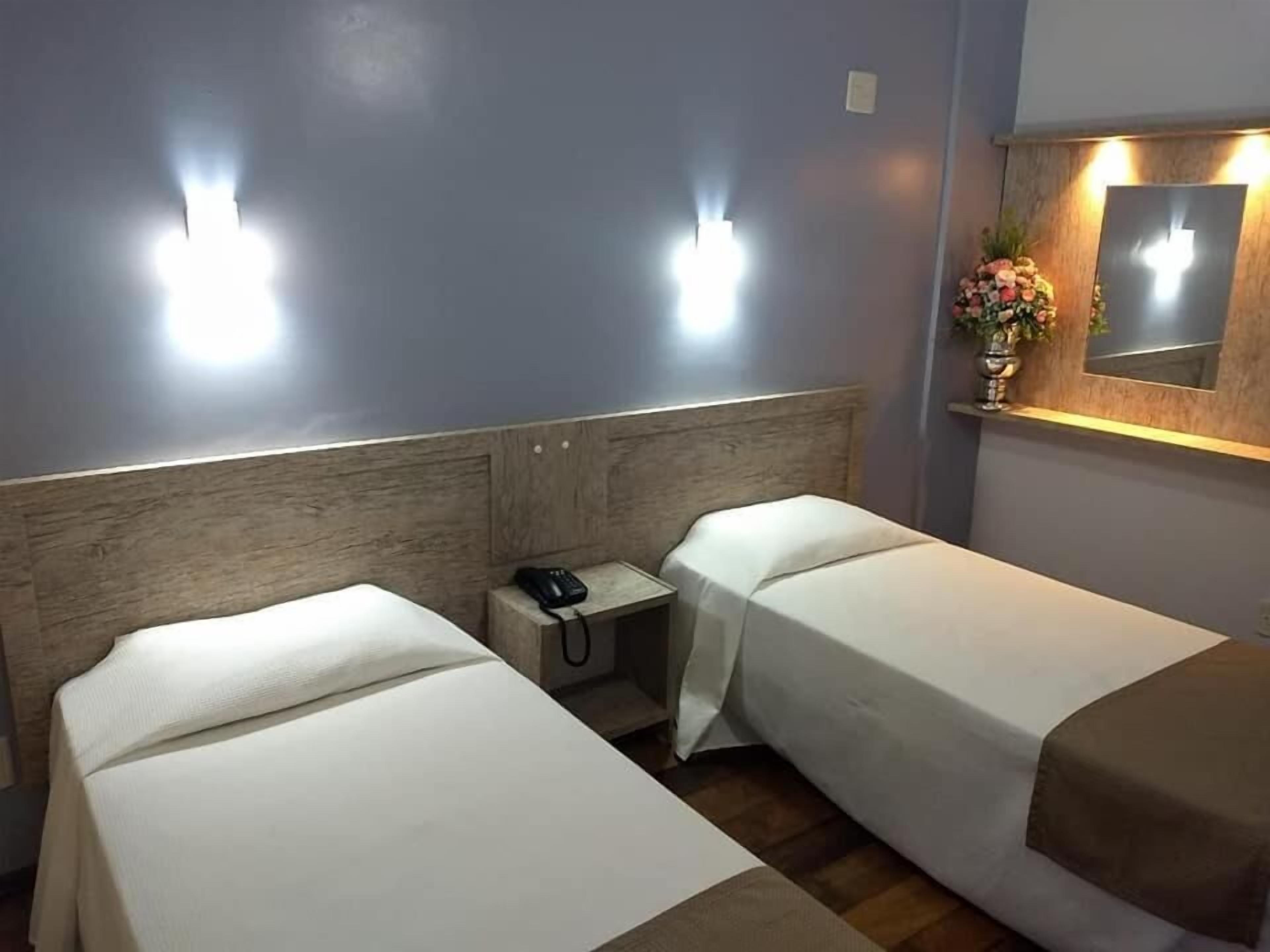 Quarto de hotel com duas camas de solteiro, uma mesa de cabeceira com telefone e um espelho sobre a cama.