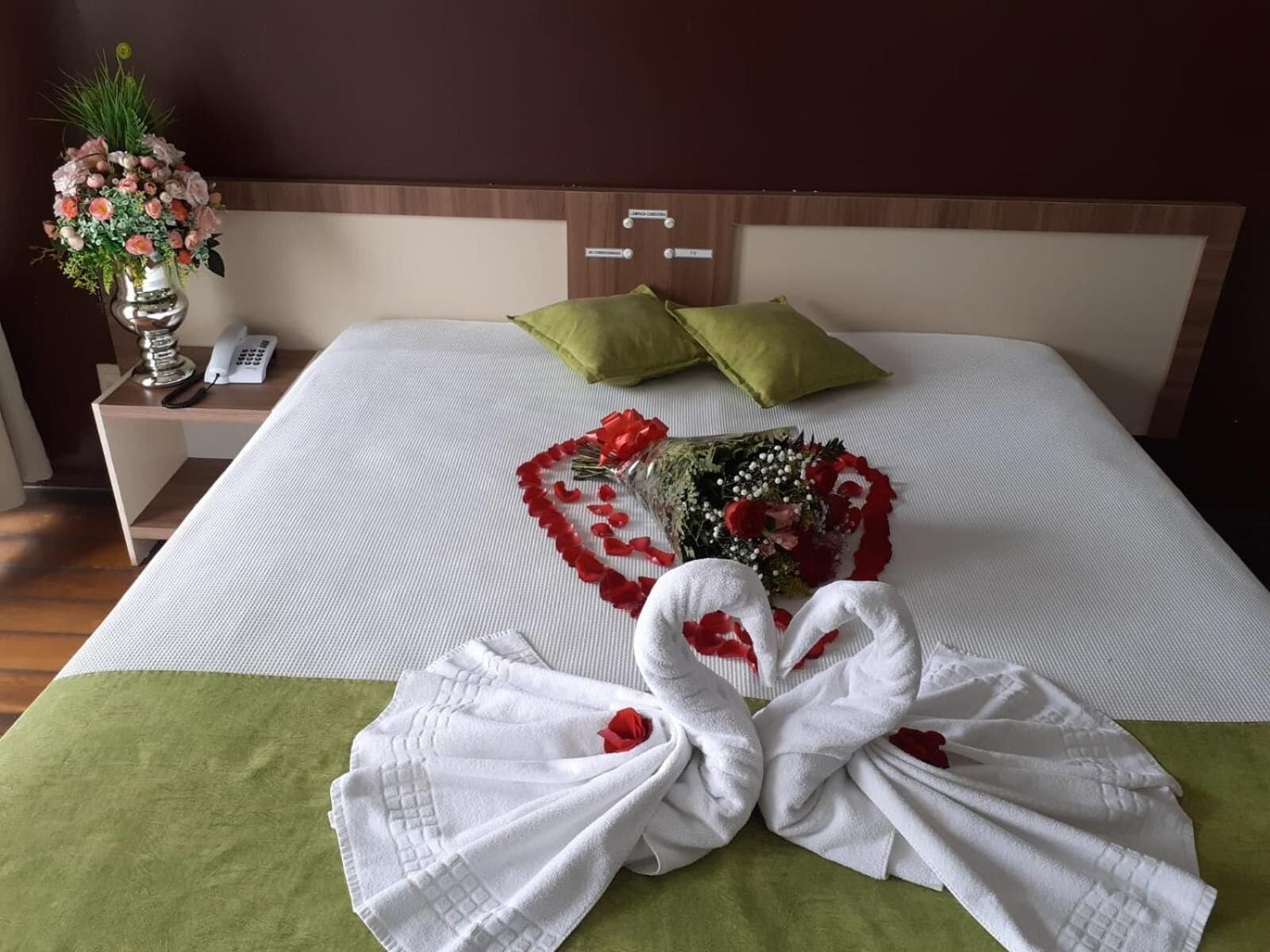 A hotel room with a neatly made bed, a bouquet of flowers, and a bouquet of rose petals arranged in a heart shape.