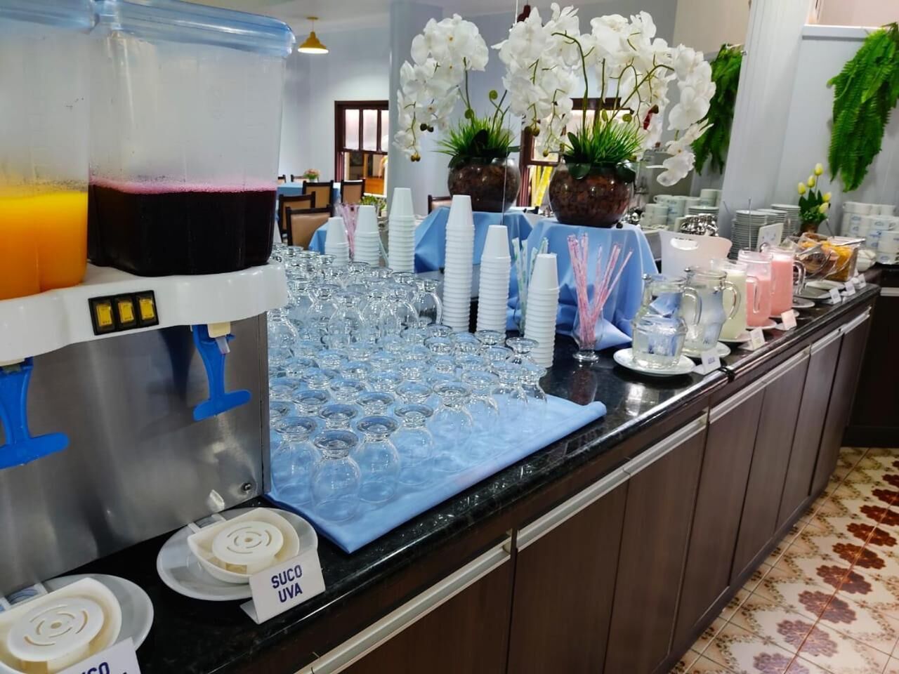 Disposição de buffet com dispensador de bebidas, pratos empilhados e diversos tipos de copos.