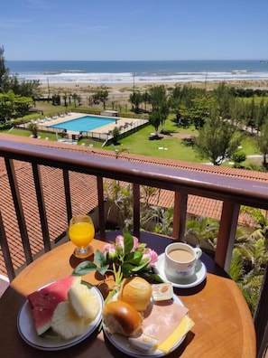 Un balcón con una mesa lista para el desayuno, con vista a una piscina y una playa.