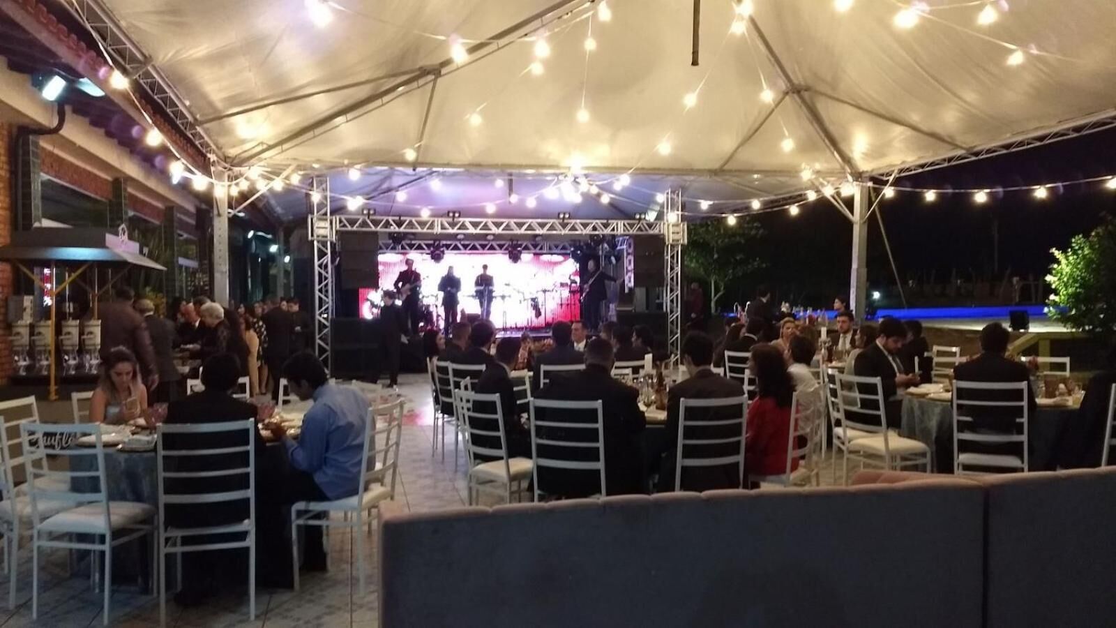 Uma área de refeições ao ar livre com mesas e cadeiras sob uma tenda, um palco com artistas e uma piscina ao fundo.