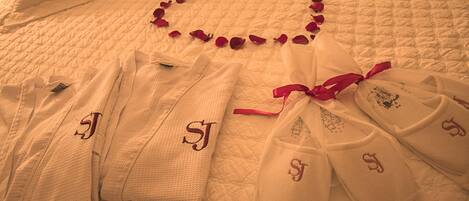 A bed with a heart-shaped arrangement of rose petals, neatly folded white robes with embroidered initials, and a red ribbon tied around one robe.