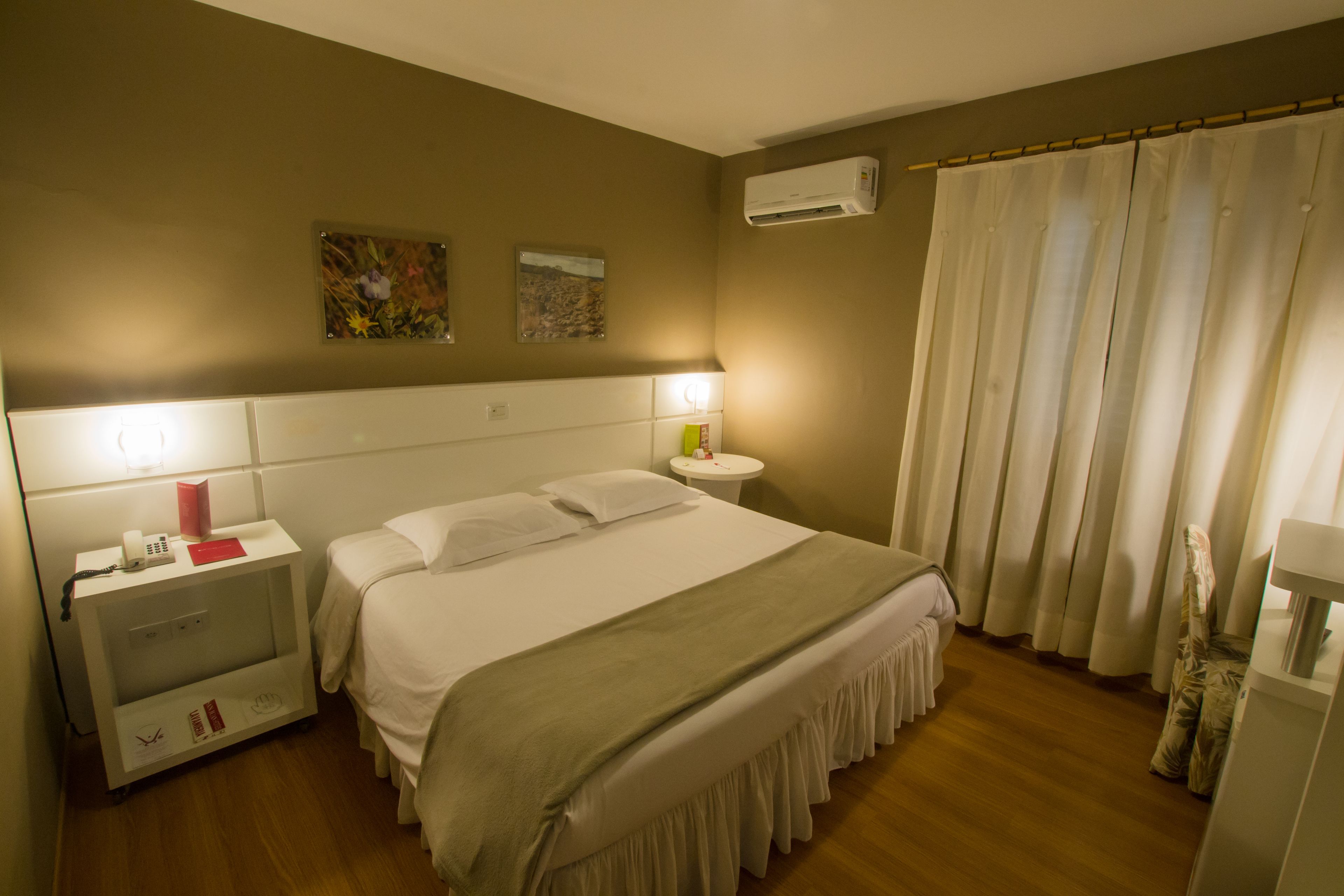 Une chambre d’hôtel comprenant un lit, des tables de chevet, une petite table, un climatiseur fixé au mur et un rideau.