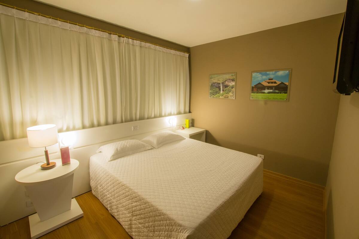 Un lit bien fait, avec une courtepointe, une lampe de chevet et une table de chevet sur laquelle se trouvent une lampe et un objet rose.