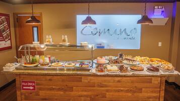 Ein Buffet mit verschiedenen Desserts und Getränken, ein Schild, das auf einen besonderen Raum hinweist, und eine Projektionsleinwand mit dem Wort "Comino".