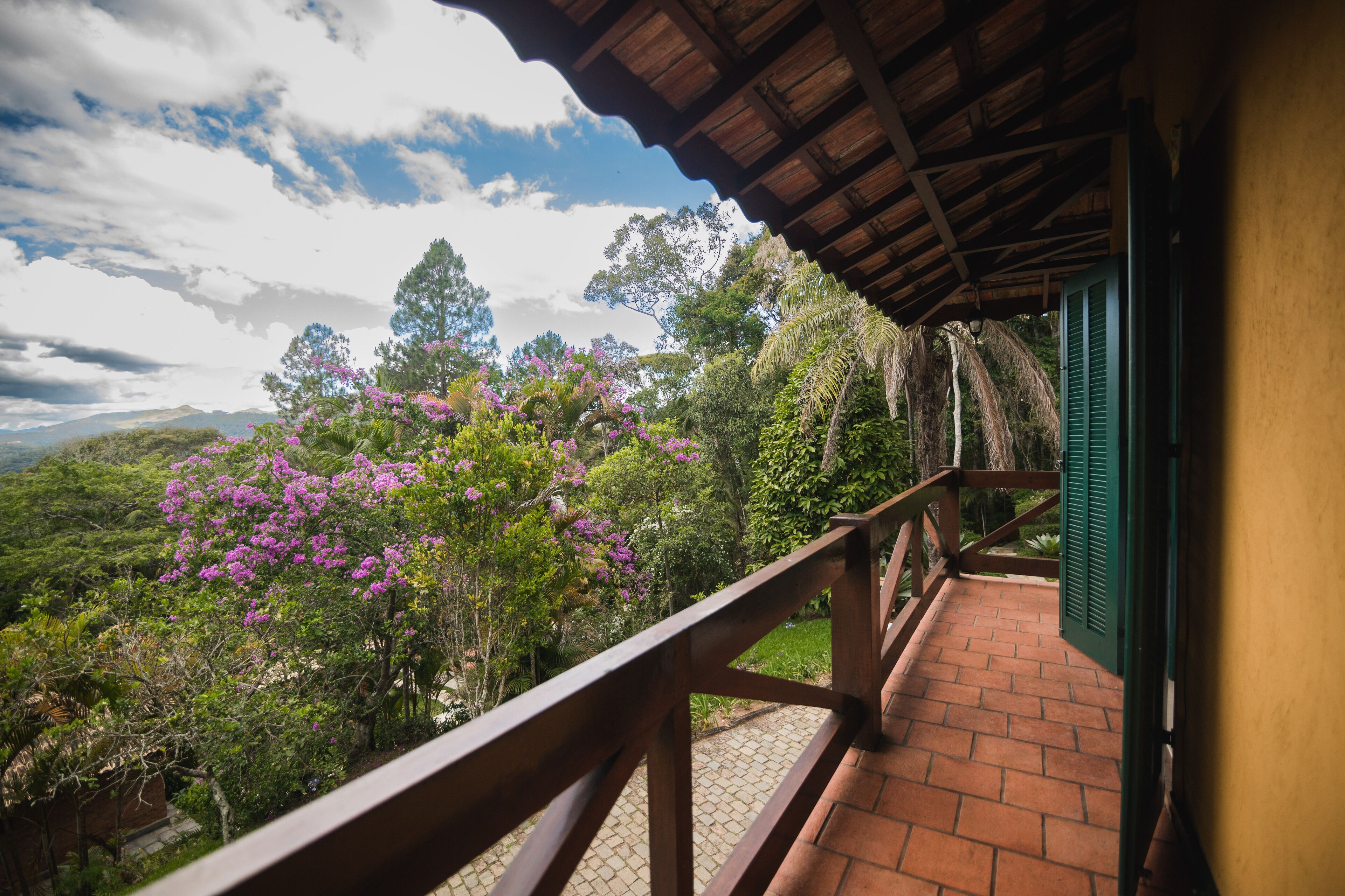 Un balcón con baranda de madera que da a un jardín exuberante con flores moradas y vistas a montañas distantes.