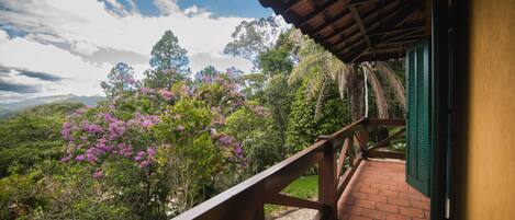 Un balcón con baranda de madera que da a un jardín exuberante con flores moradas y vistas a montañas distantes.