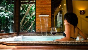 Una persona relajándose en un jacuzzi con una copa de vino tinto, rodeada de un muro de ladrillo y grandes ventanales con vistas a la vegetación.