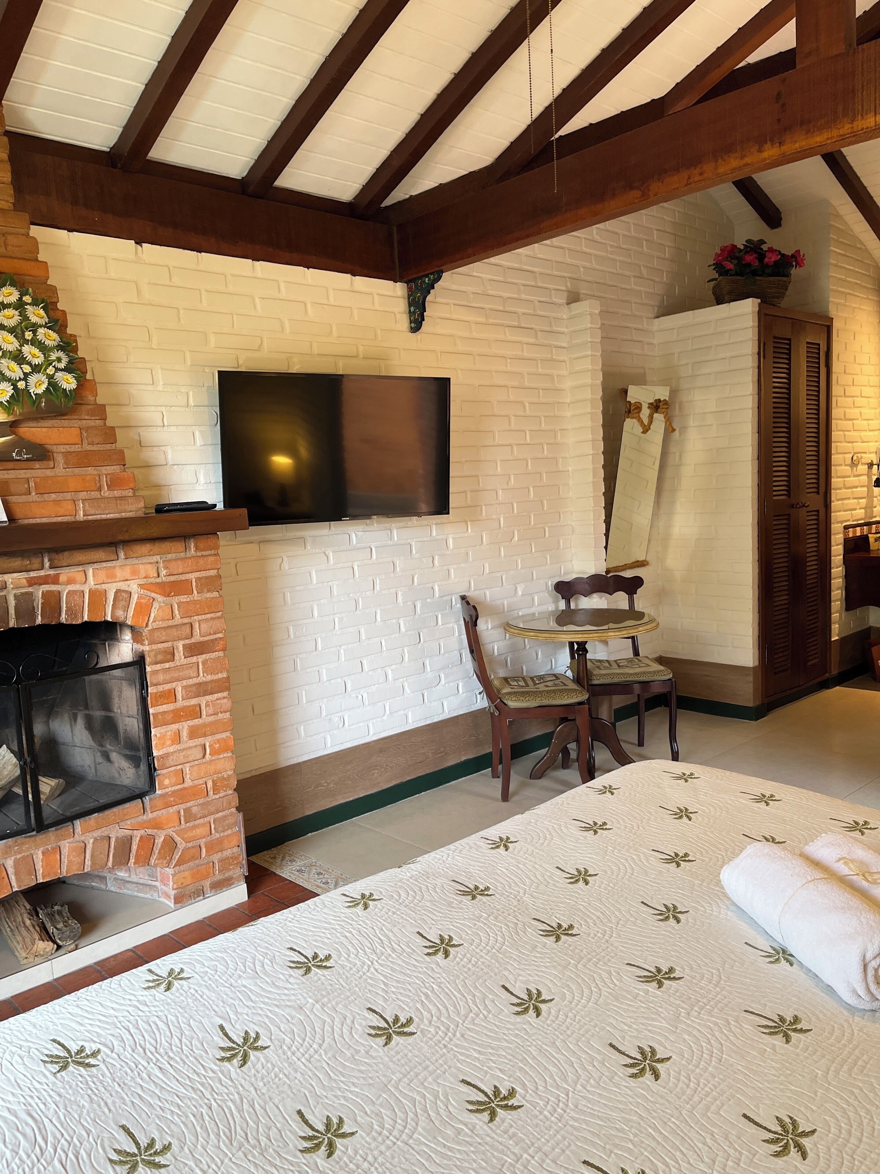 Habitación acogedora con chimenea de ladrillo, un televisor de pantalla plana montado en una pared de ladrillo blanco, una silla de madera y una cama con colcha estampada.