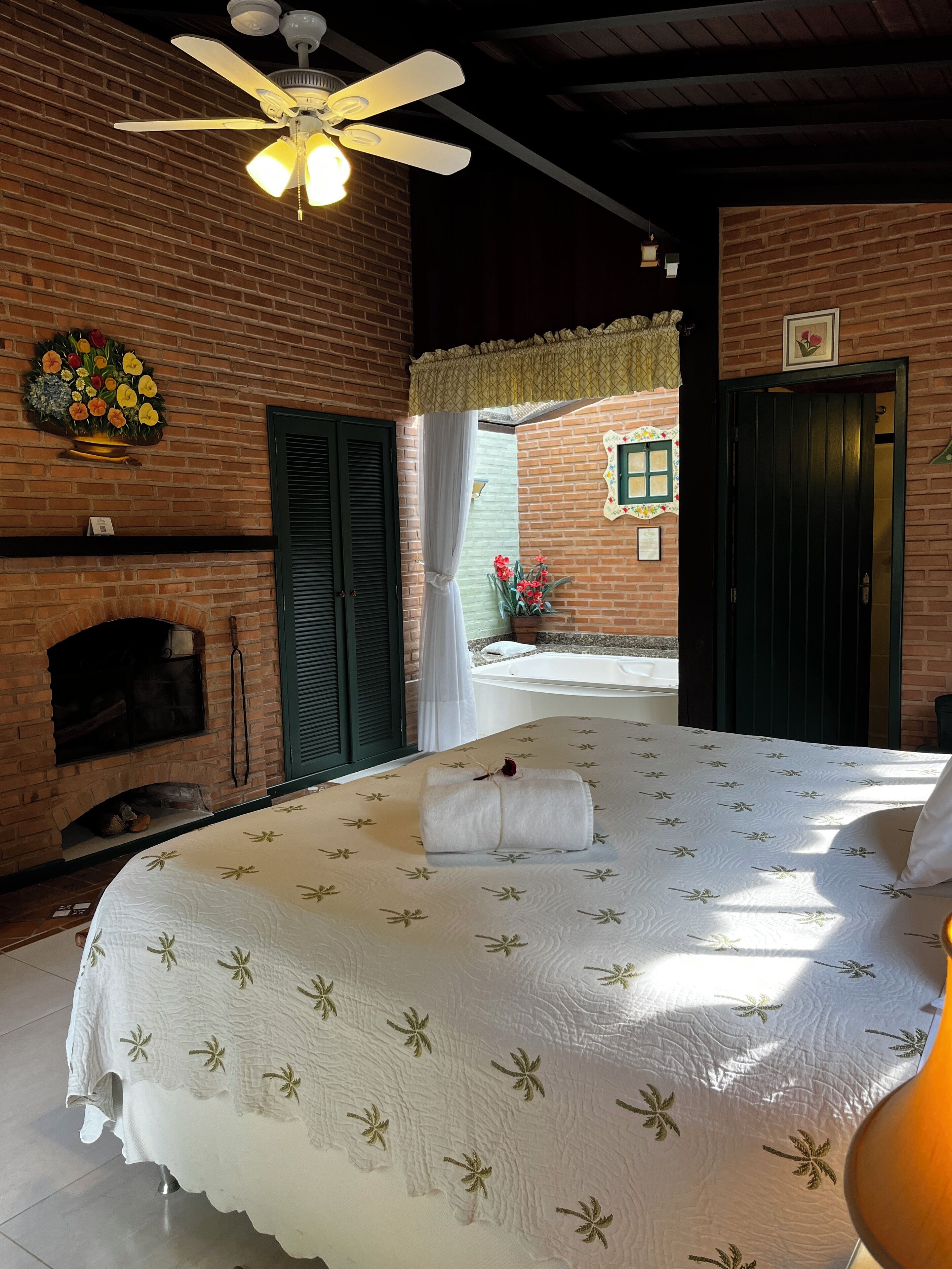 A bedroom with a brick wall, a ceiling fan, a fireplace, a bed with a floral bedspread, and a view of an outdoor area.