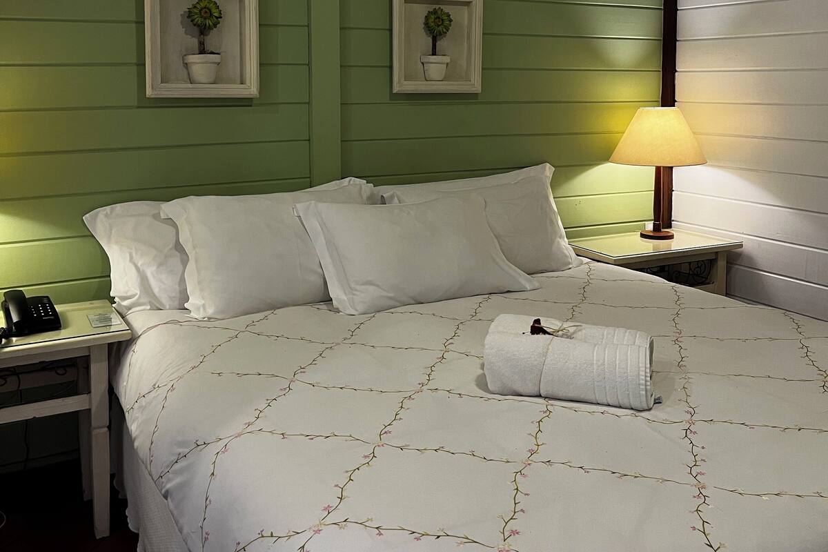 Een net opgemaakt bed met witte lakens en een bloemenmotief, een bedlampje, een nachtkastje met een telefoon en twee ingekorte plantenpotten aan de muur.