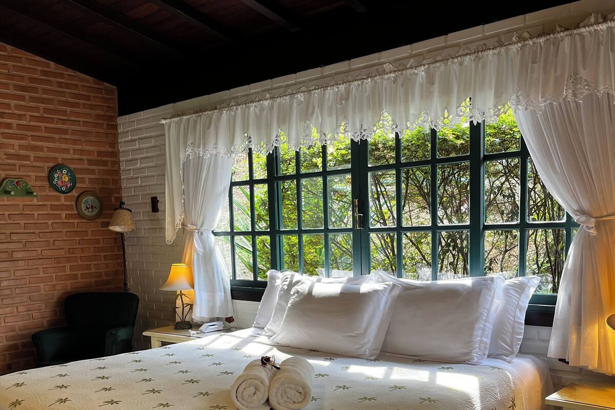 Een slaapkamer met een groot bed, een bankje, een nachtkastje en uitzicht op groen door een raam.