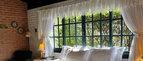A bedroom with a large bed, a bench, a nightstand, and a view of greenery through a window.