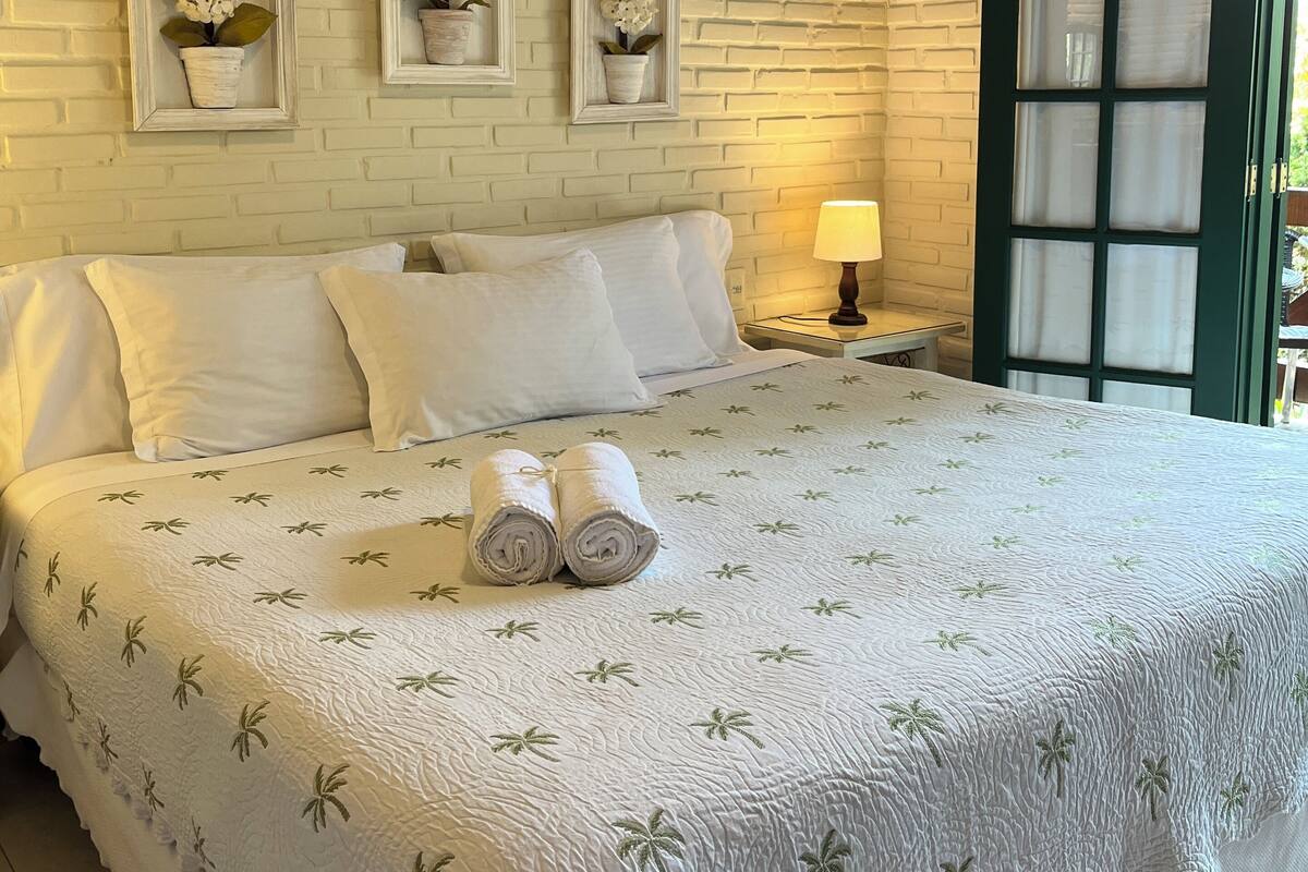 Een netjes opgemaakt bed met een witte spreidek die palmboommotieven heeft, een houten hoofdbord en een muur met een baksteenpatroon, versierd met drie planten in potten.