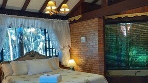 Un dormitorio con cama, ventana con cortinas, pared de ladrillo y mesita de noche con lámpara.