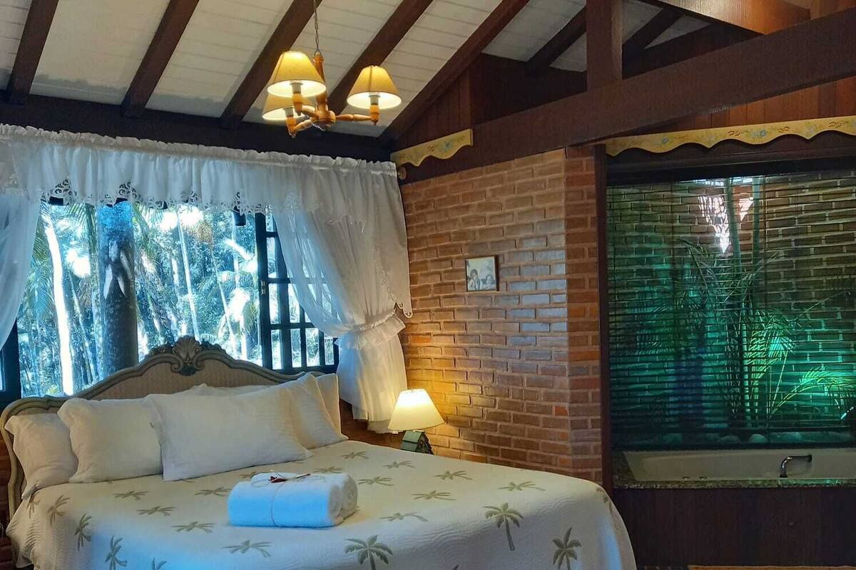 Een slaapkamer met een bed, een raam met gordijnen, een bakstenen muur en een nachtkastje met een lamp.