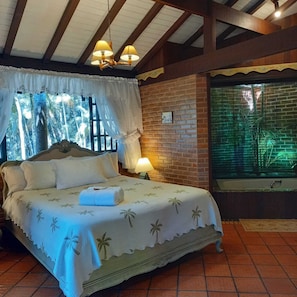 Een slaapkamer met een bed, een raam met gordijnen, een bakstenen muur en een nachtkastje met een lamp.