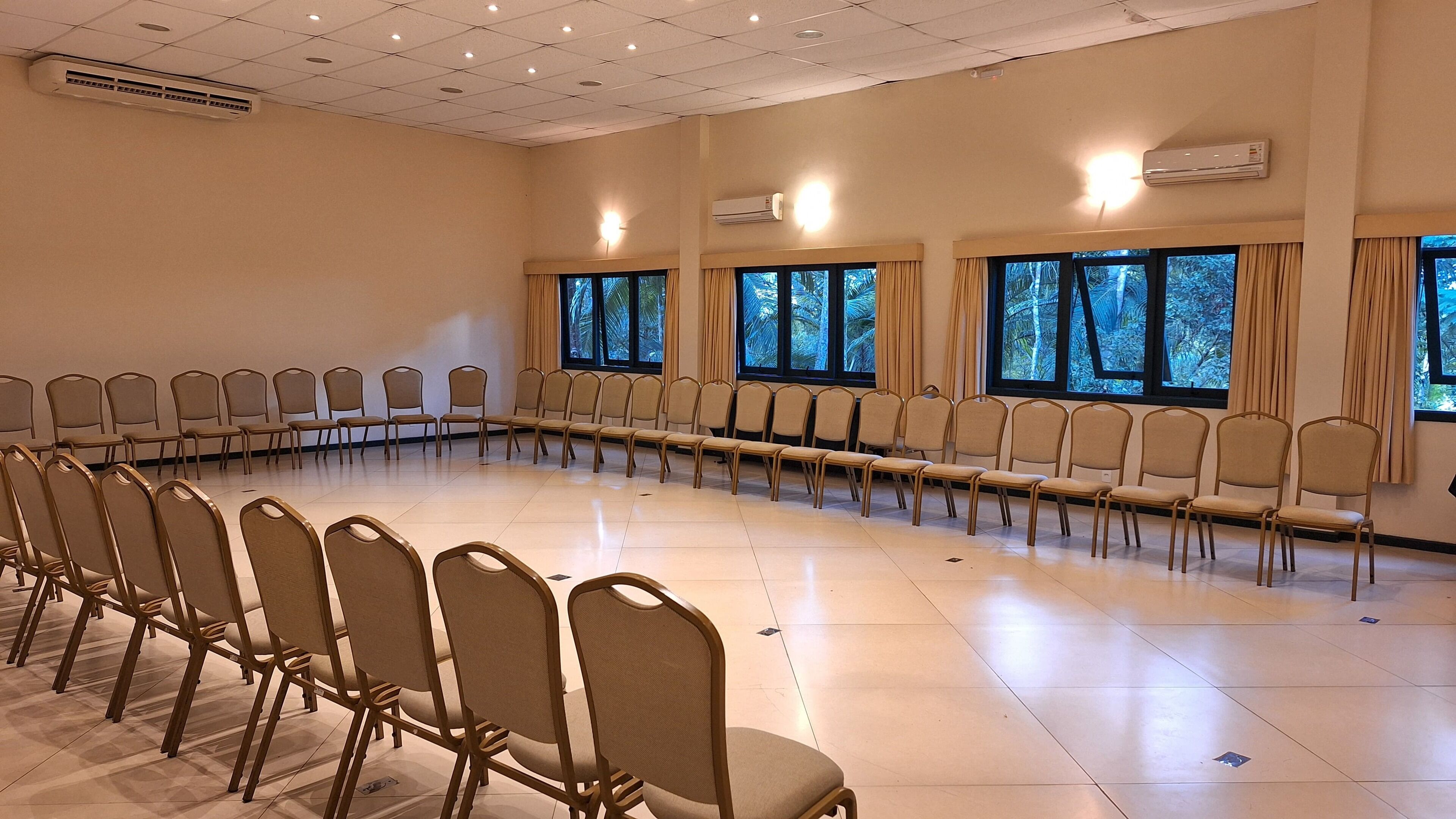 A conference room with rows of chairs, large windows with curtains, and ceiling lights.