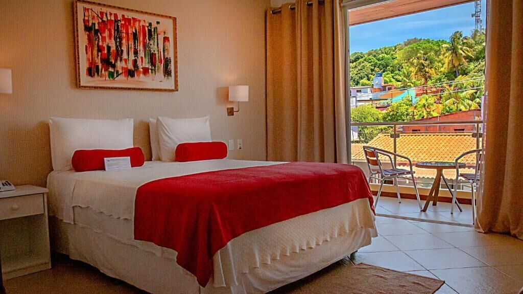 A hotel room with a bed, a nightstand, a window with curtains, and a balcony with a table and chairs.