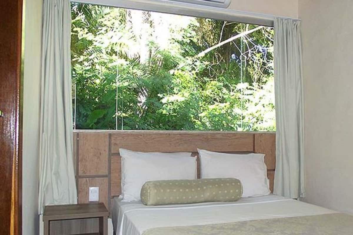 Un dormitorio con cabecera de madera, cama con ropa de cama blanca y vista a la vegetación a través de una ventana.