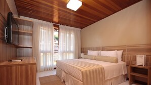 Un dormitorio con techo de madera, cama con ropa blanca, mesita de noche, televisor montado en la pared y ventana con cortinas.