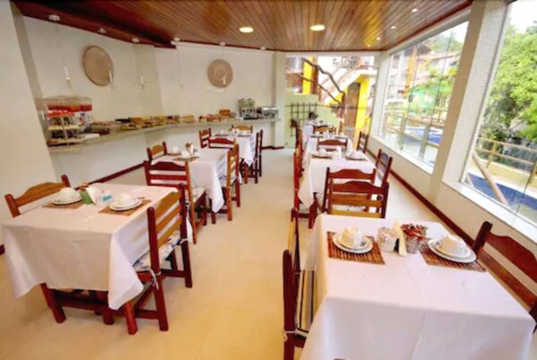 A dining area with multiple tables and chairs, a counter with food items, and large windows offering a view of the outdoors.