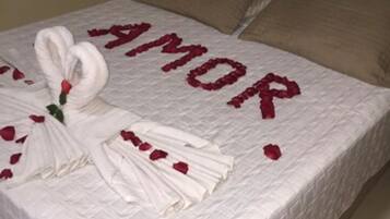 A bed with a white bedspread, a towel arrangement spelling "AMOR", and rose petals.