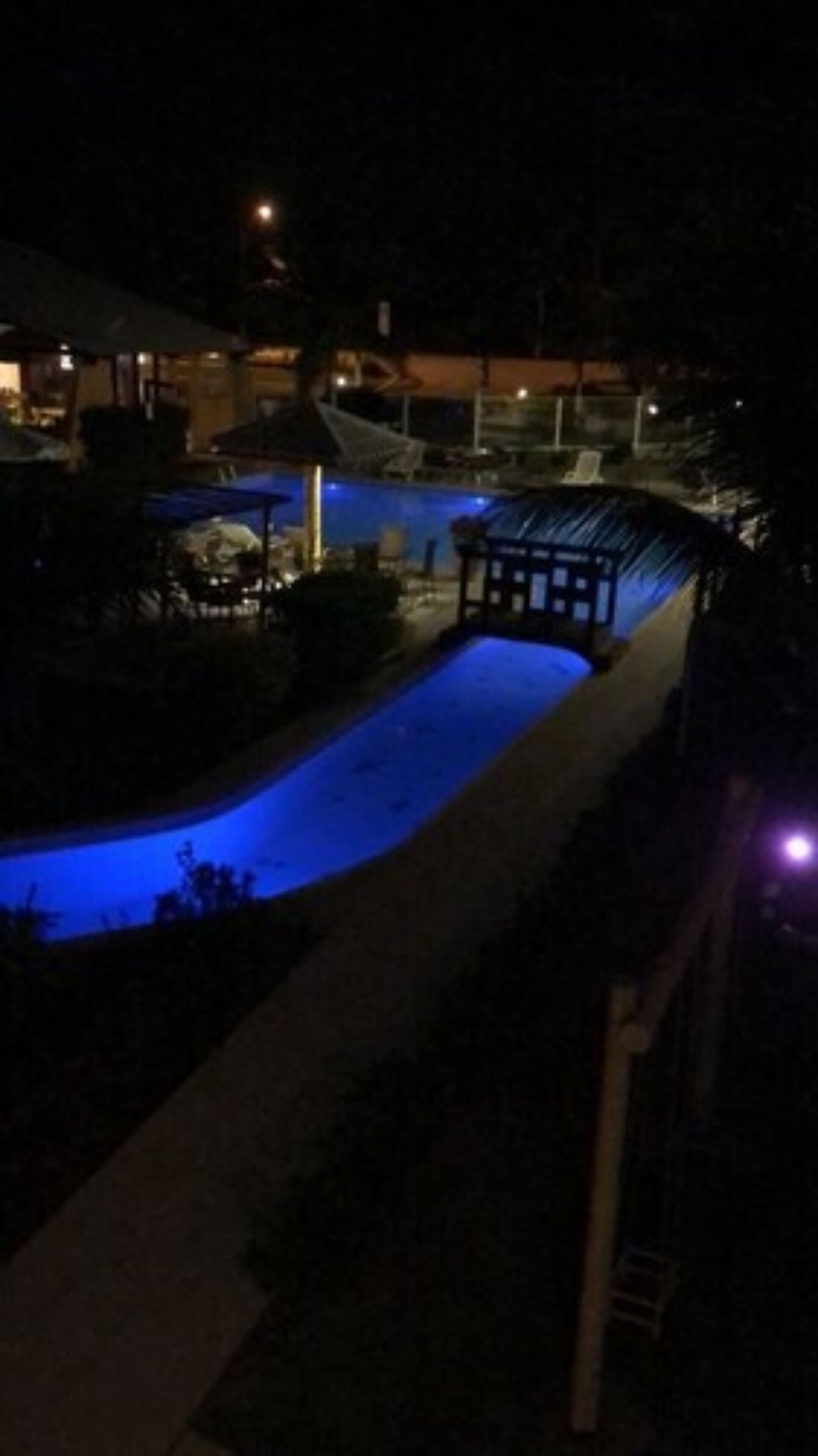 A nighttime view of a swimming pool area with a covered seating area and a building in the background.
