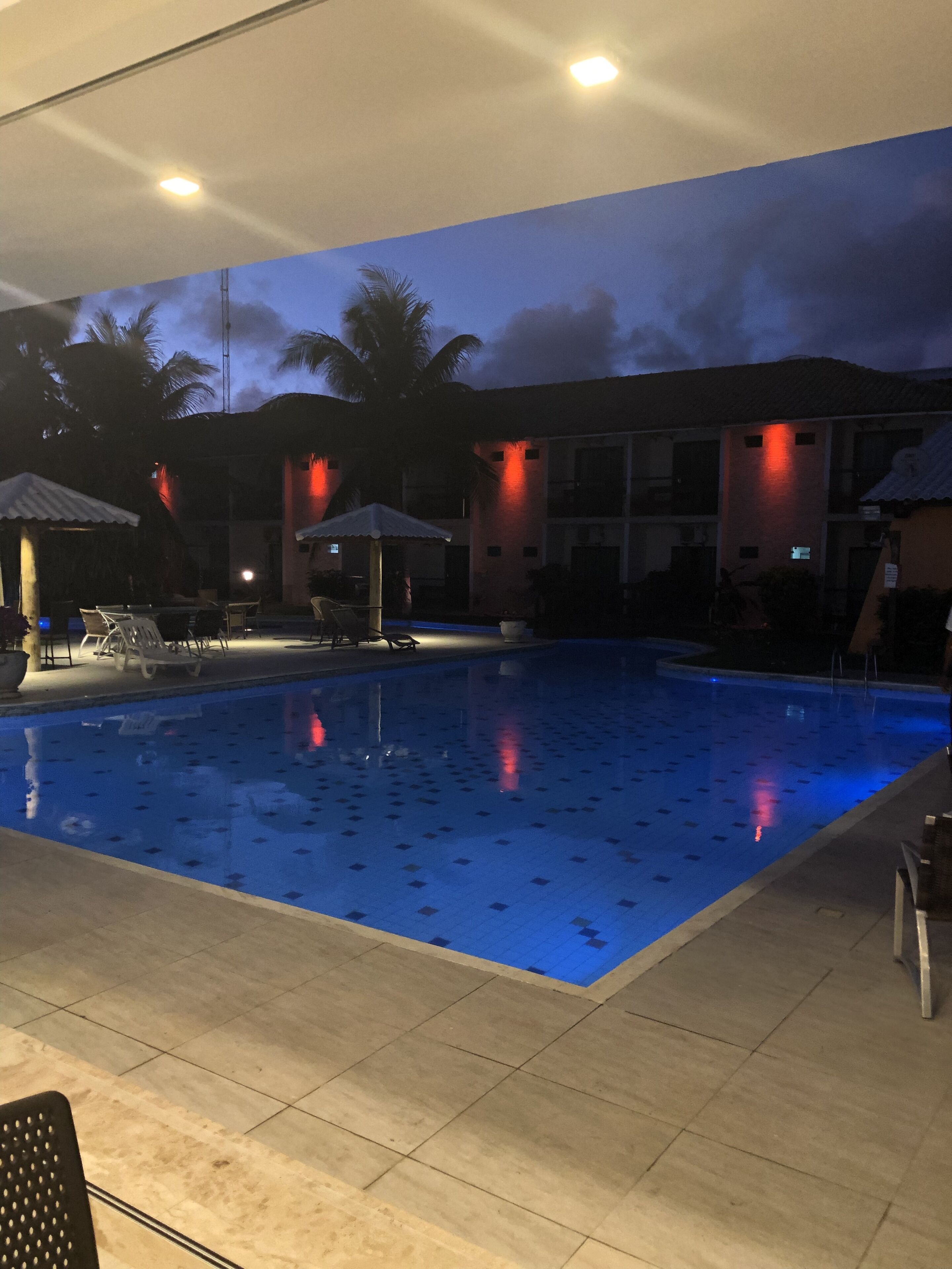 A swimming pool with tiled flooring, lounge chairs, and umbrellas.
