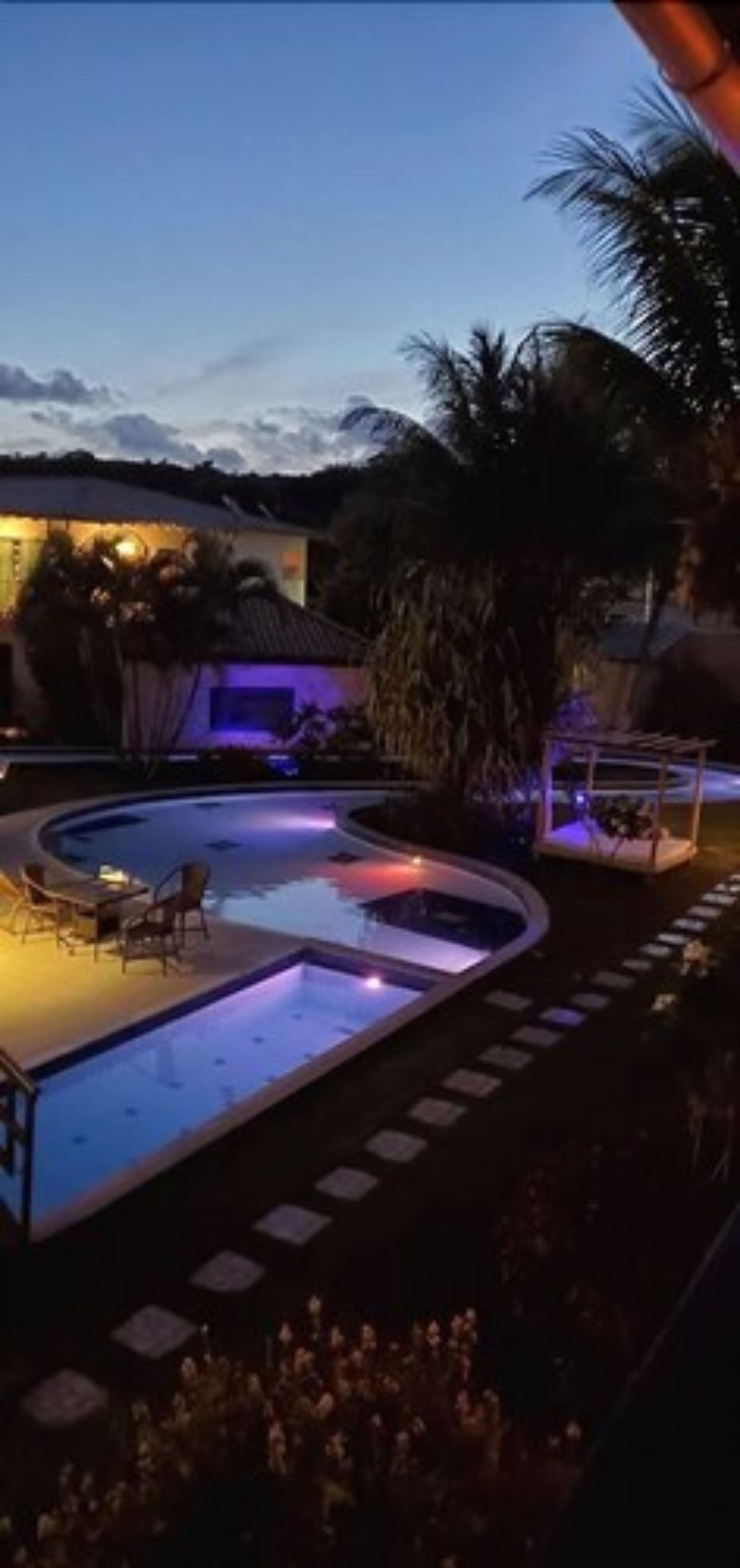 Ein Poolbereich mit Liegestuhl und Tisch, umgeben von Palmen und einem Gebäude mit einem Reetdach.