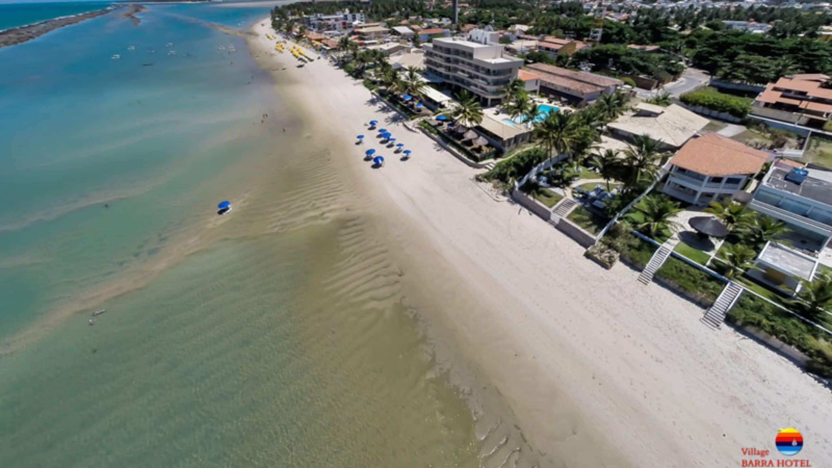 Vista aérea de uma praia com um hotel chamado Village Barra Hotel. A água é cristalina e a orla da praia está repleta de guarda-sóis e espreguiçadeiras.