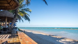 Une terrasse en bois agrémentée de parasols en chaume, une plage de sable et une mer d’un bleu limpide.