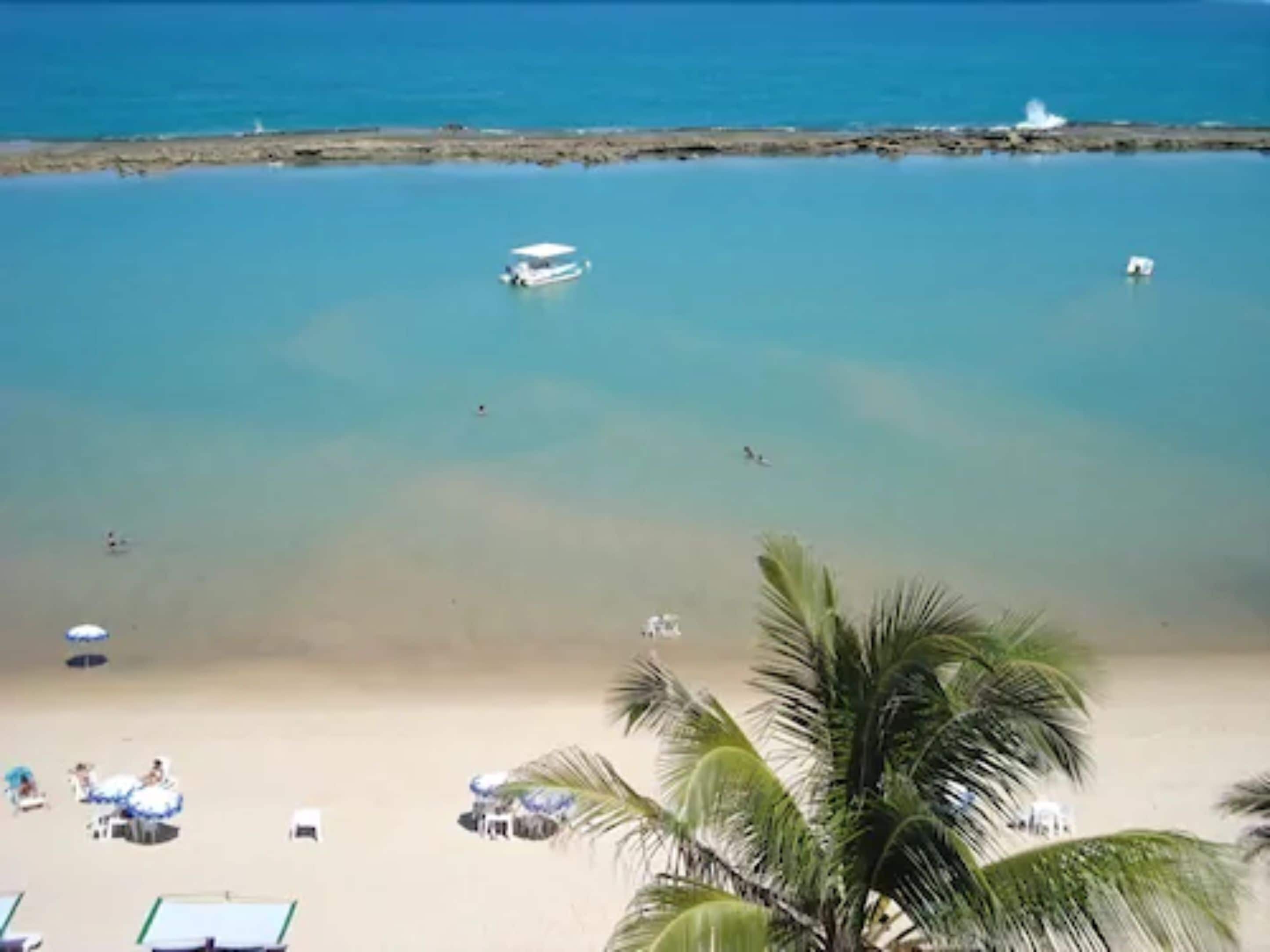A beach with clear blue water, a few boats, and palm trees.