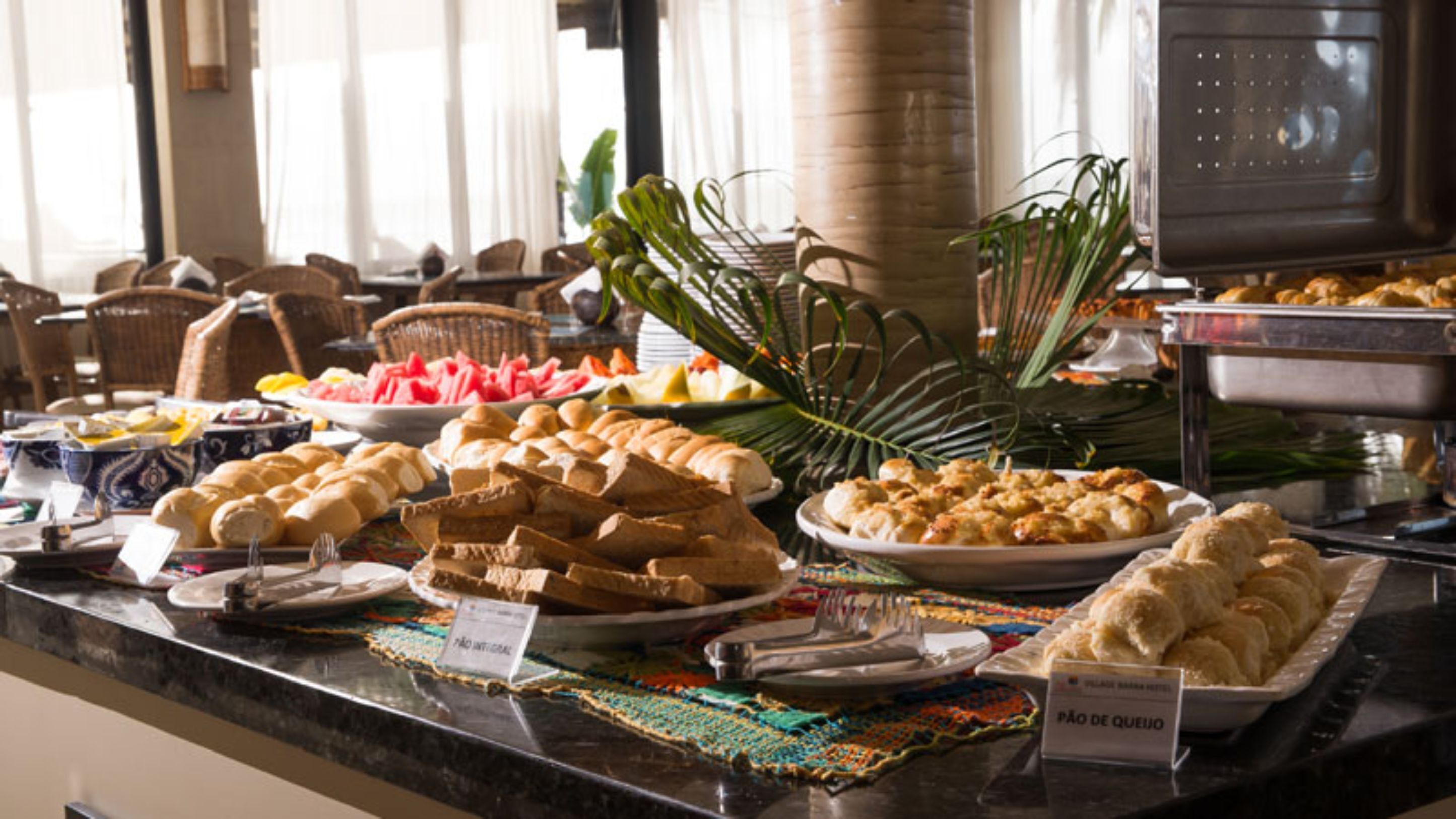 A buffet with various food items including bread, pastries, and a plate labeled "Pão de Queijo".