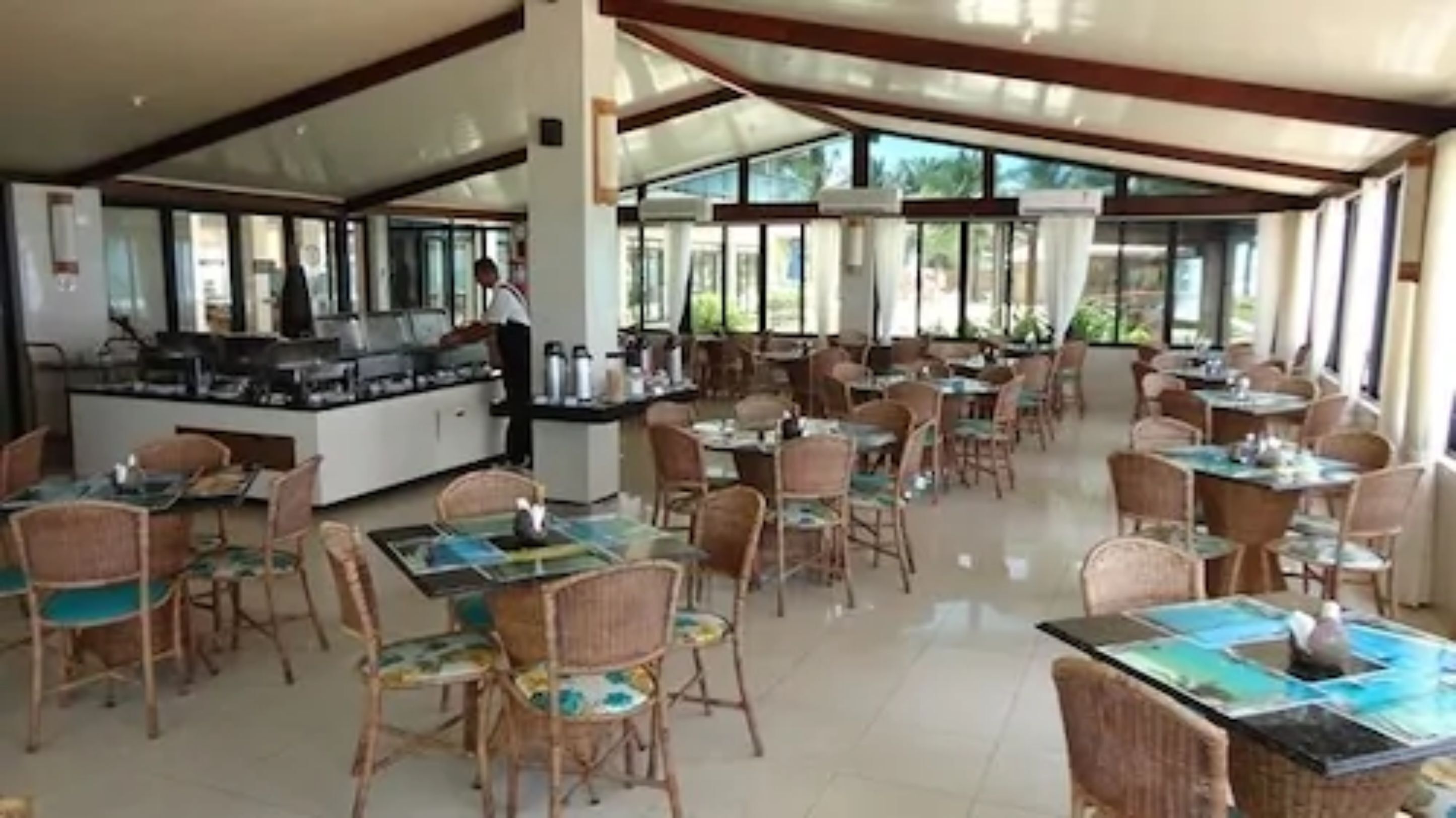 A spacious restaurant with multiple tables and chairs, a central counter with a person serving, and large windows allowing natural light.