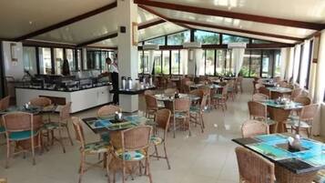 A spacious restaurant with multiple tables and chairs, a central counter with a person serving, and large windows allowing natural light.