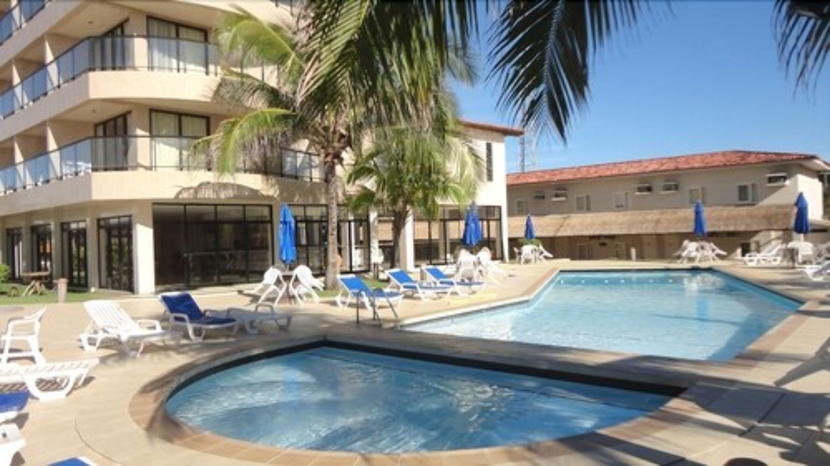 A swimming pool area with lounge chairs and umbrellas, surrounded by buildings with balconies and palm trees.