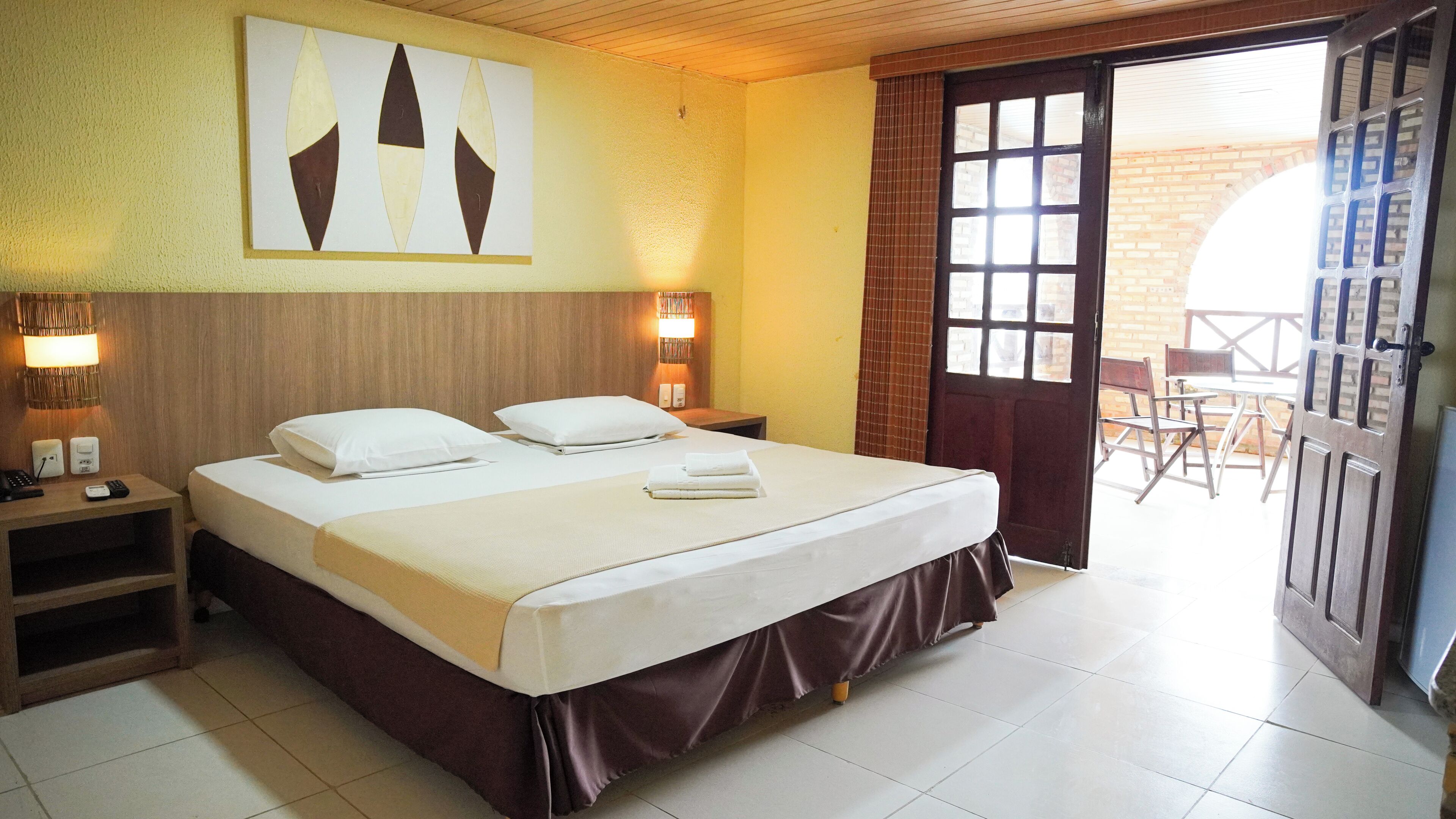 Habitación de hotel con cama, lámparas de noche, una obra de arte colgada en la pared, y una puerta que da a un espacio exterior con mesa y sillas.