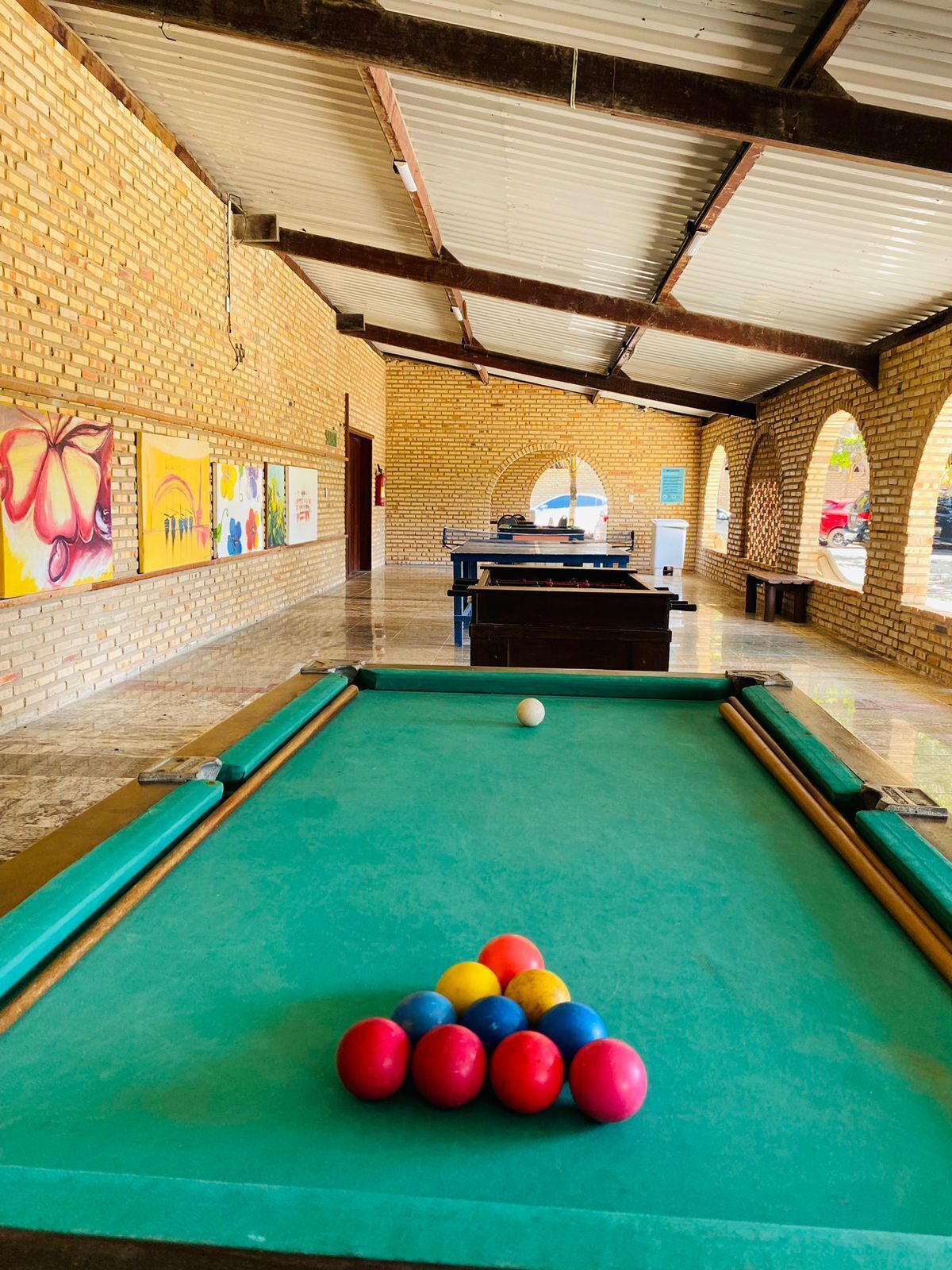A billiards table with balls arranged in a pyramid, a pool table, and a brick wall with paintings.