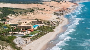Vista aérea de un complejo turístico costero con playa, océano y aerogeneradores al fondo.