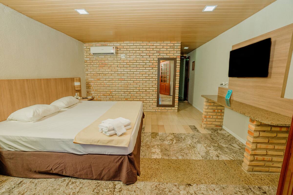 A hotel room with a large bed, a wooden headboard, a brick accent wall, a flat-screen TV mounted on the wall, and a small desk with a mirror.
