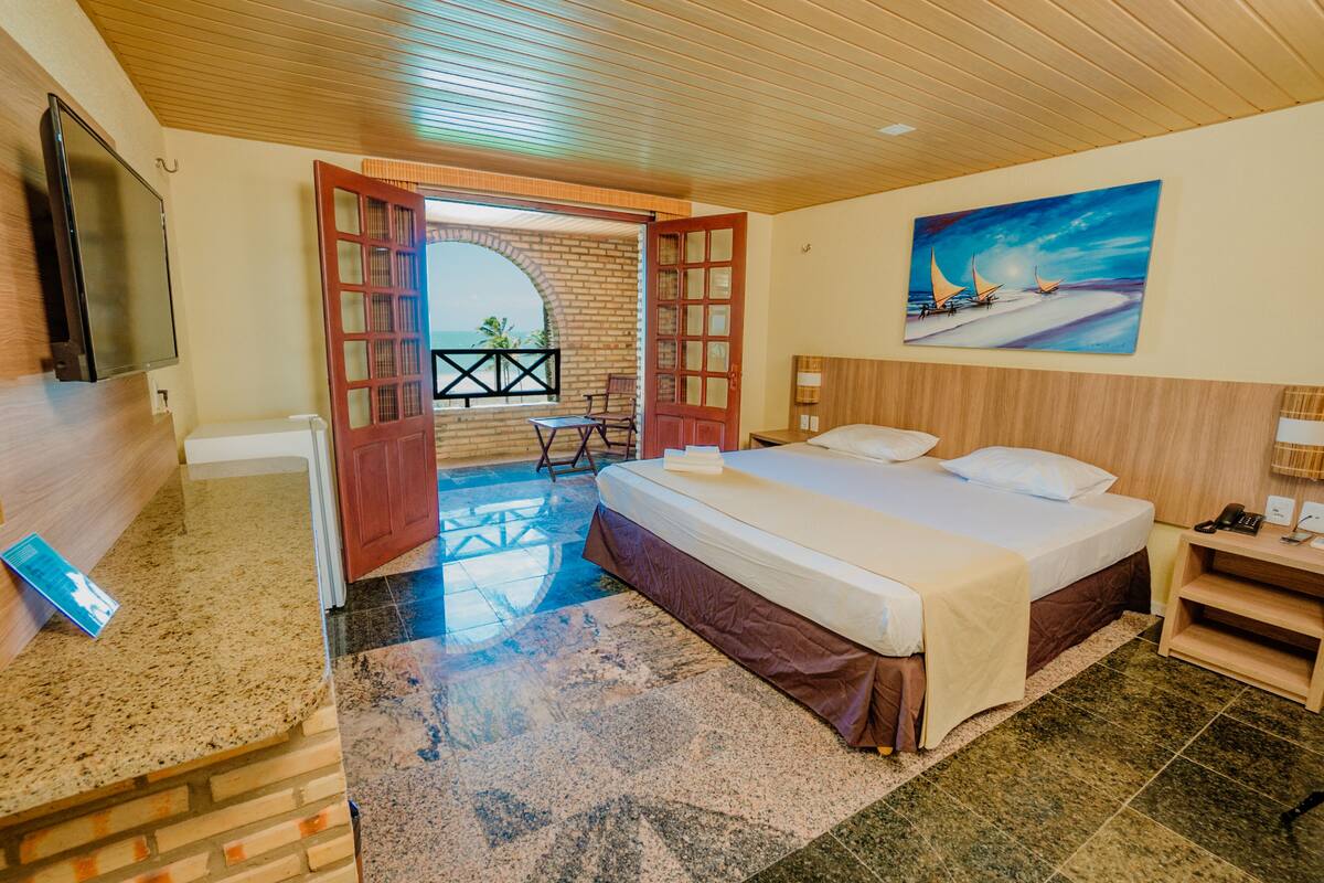 A hotel room with a bed, a TV, a painting, and a view of the beach.