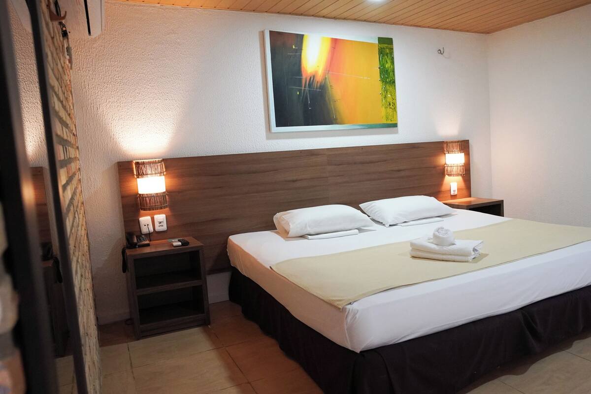 A hotel room with a bed, two bedside lamps, a nightstand, a wall-mounted painting, and a wooden ceiling.
