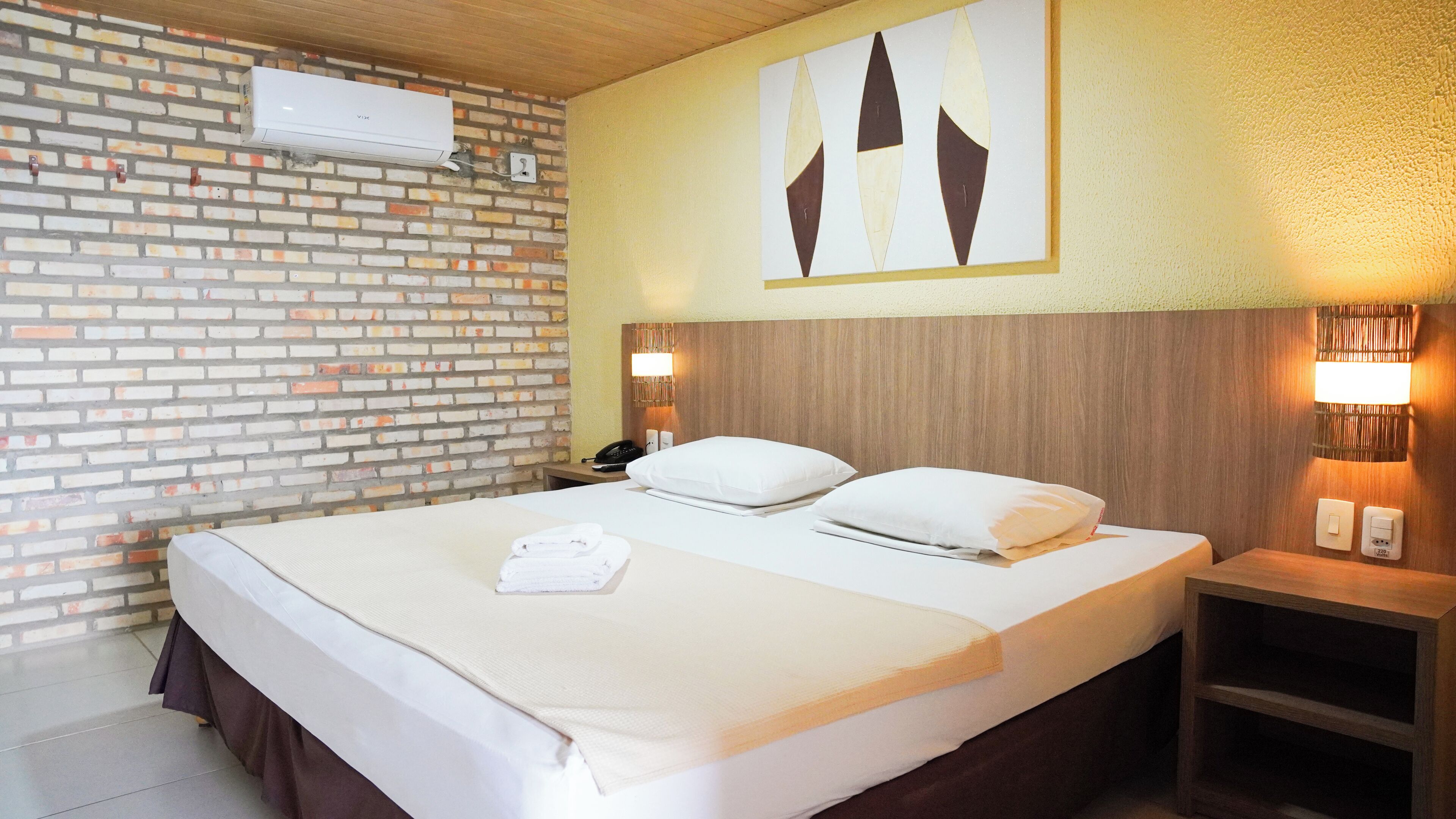 A hotel room with a brick-patterned wall, a bed with white linens, a bedside table with a phone, and a wall-mounted light fixture.