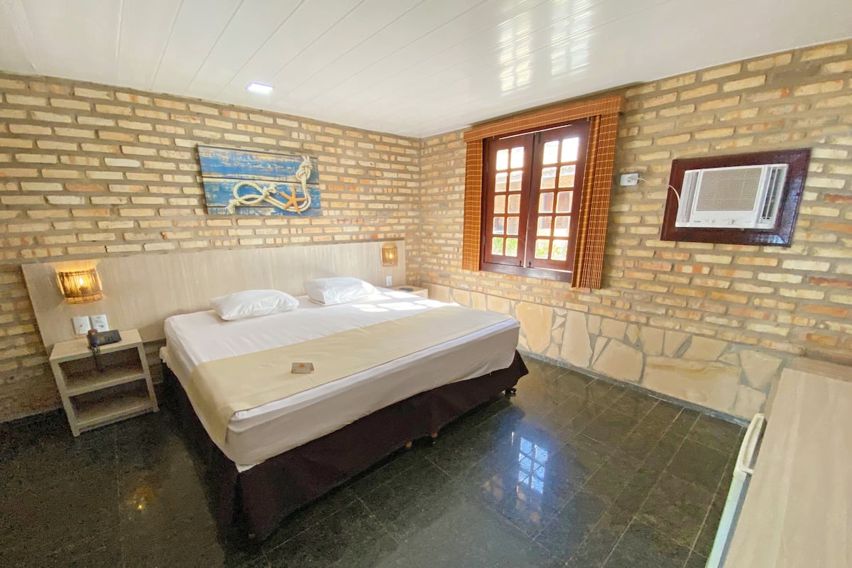 A bedroom with a brick wall, a bed with white linens, a nightstand, a wall-mounted air conditioner, and a window with brown shutters.