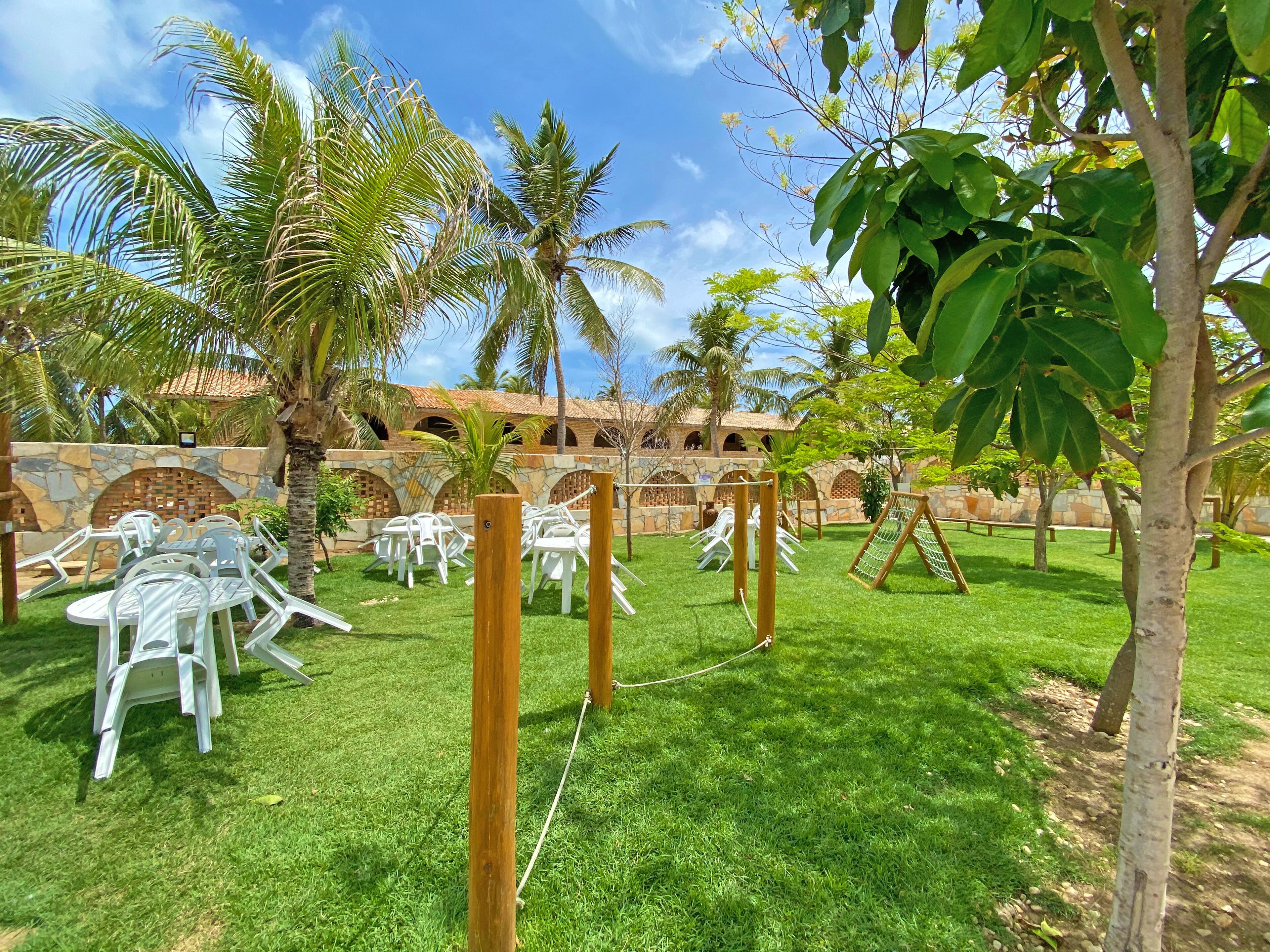 A courtyard with white plastic chairs and tables, surrounded by palm trees and a stone wall.