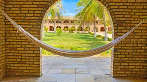 A hammock in a brick-walled room with a view of a courtyard featuring palm trees and a building with arches. - Hotel Parque das Fontes All Inclusive (Beberibe)